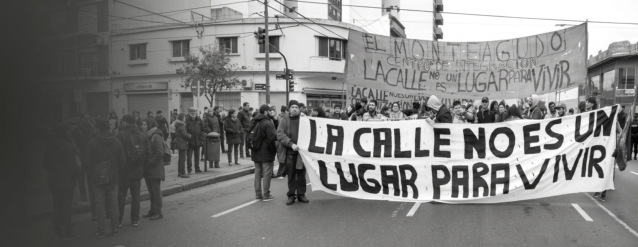 Marcha en la calle con una bandera que dice "La calle no es un lugar para vivir"; detrás, otra bandera del Centro de Integración Monteagudo.